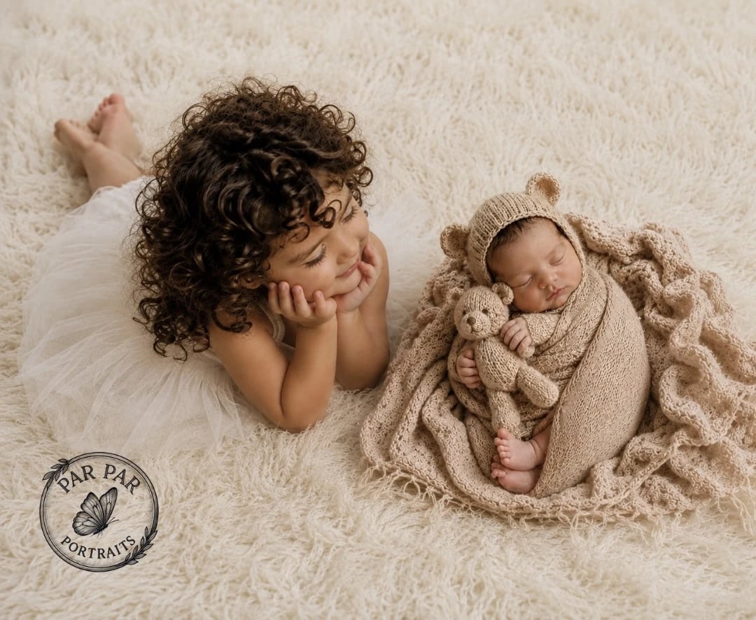 Big sister resting beside her swaddled newborn brother in a bear bonnet on a cream rug