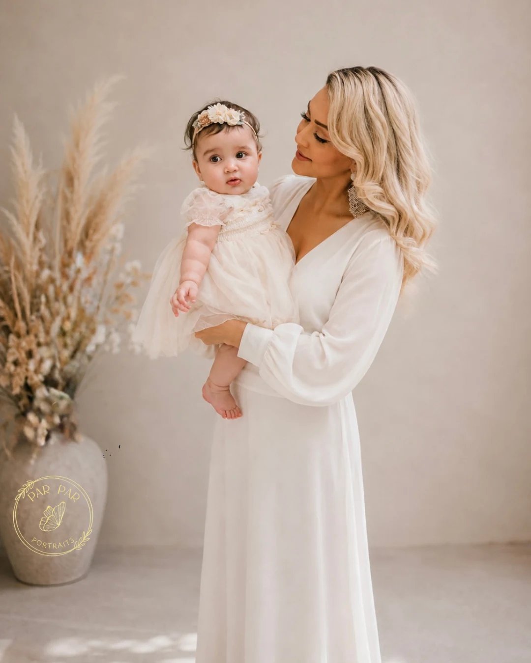 Mother in a flowing ivory gown holding her baby in a tulle dress beside dried pampas, Toronto milestone photography