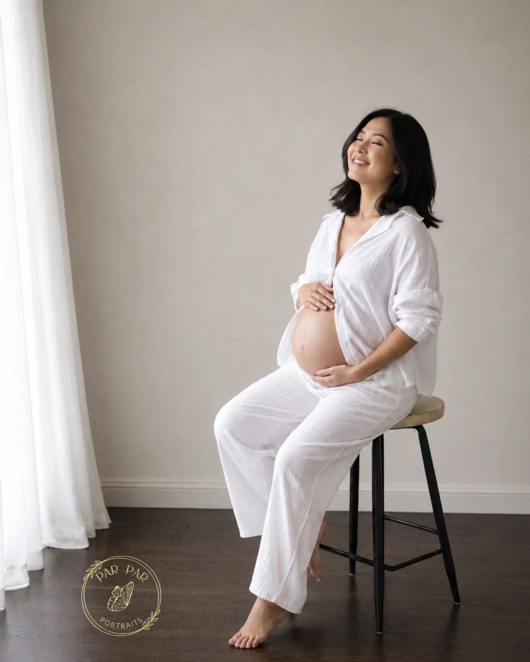 Expecting mother in white cotton laughing on a studio stool in soft daylight