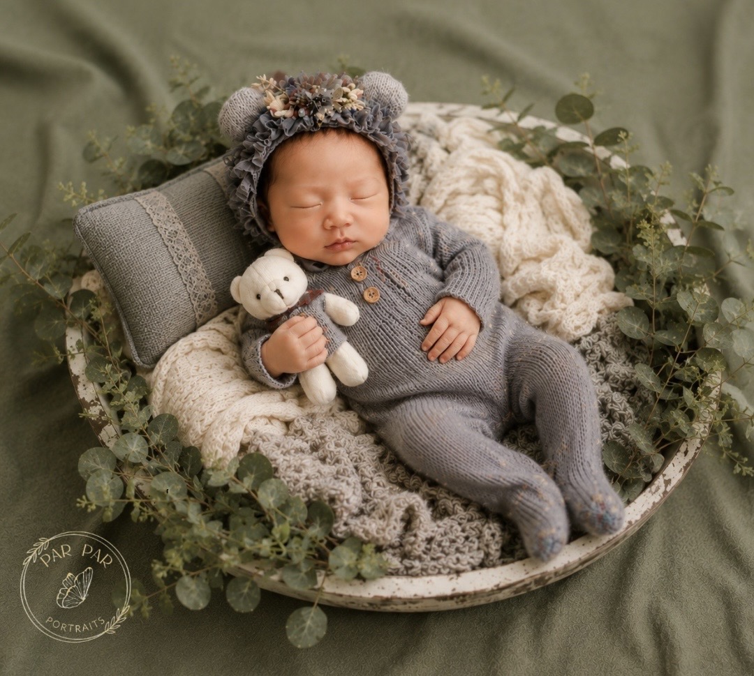 Newborn in a grey knit romper and floral bear bonnet asleep in a white bowl with eucalyptus, Toronto newborn photography