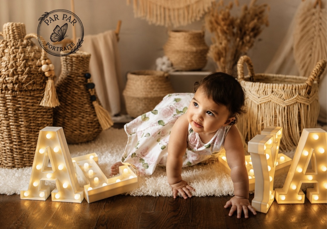 Baby girl in a rose print dress crawling beside glowing marquee name letters and woven baskets, Toronto milestone photography