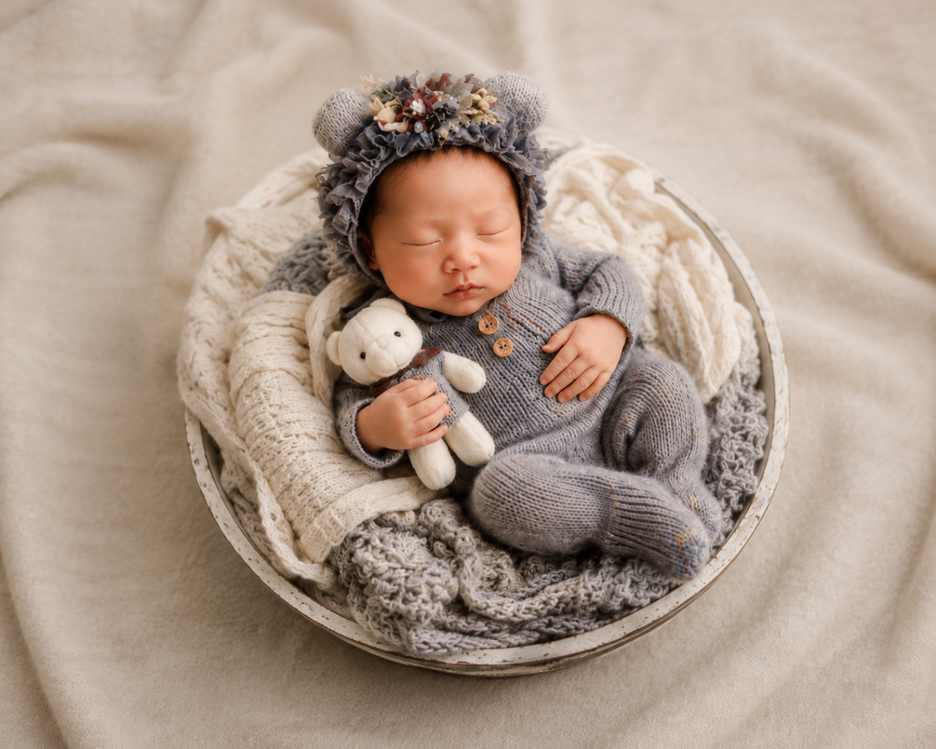 Newborn in a grey knit romper and floral bear bonnet asleep in a bowl holding a teddy