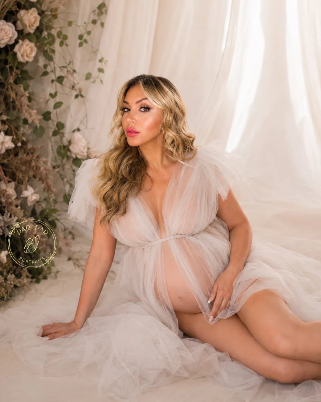 Expecting mother seated in a soft tulle gown beside a wall of cream roses and greenery