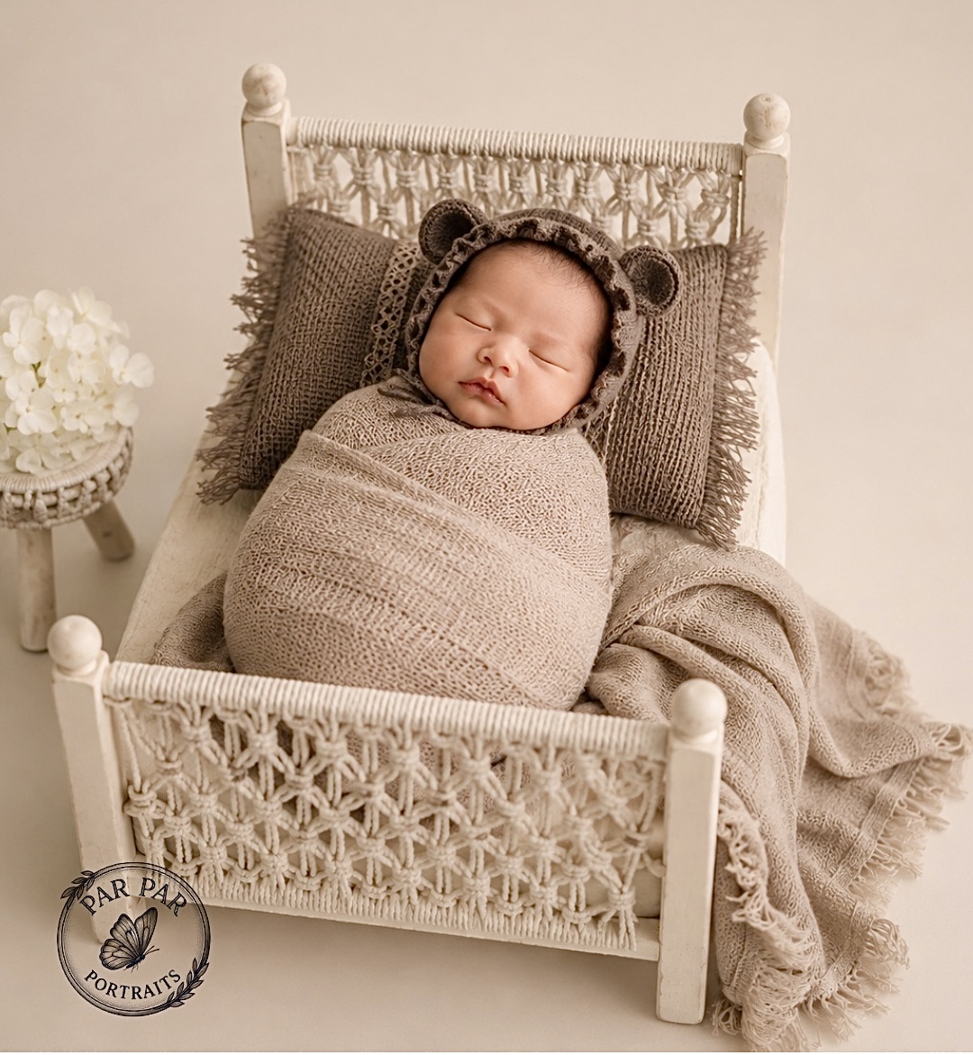 Newborn in a bear bonnet swaddled in beige on a macrame doll bed with white hydrangeas
